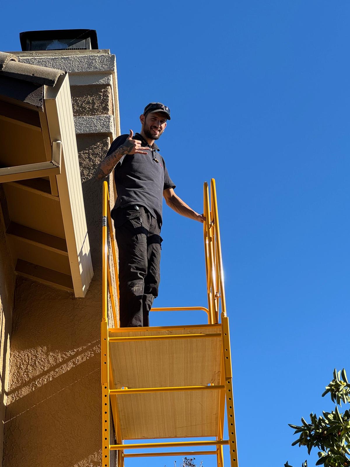 Michael standing on a yellow ladder platform beside a light stucco chimney with a dark metal chase cover