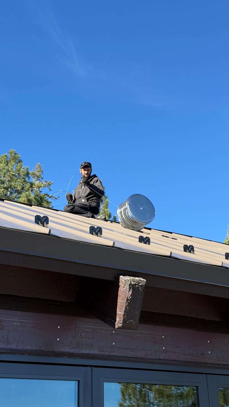 Michael seated on a standing-seam metal roof working a length of wire beside a stainless chimney cap