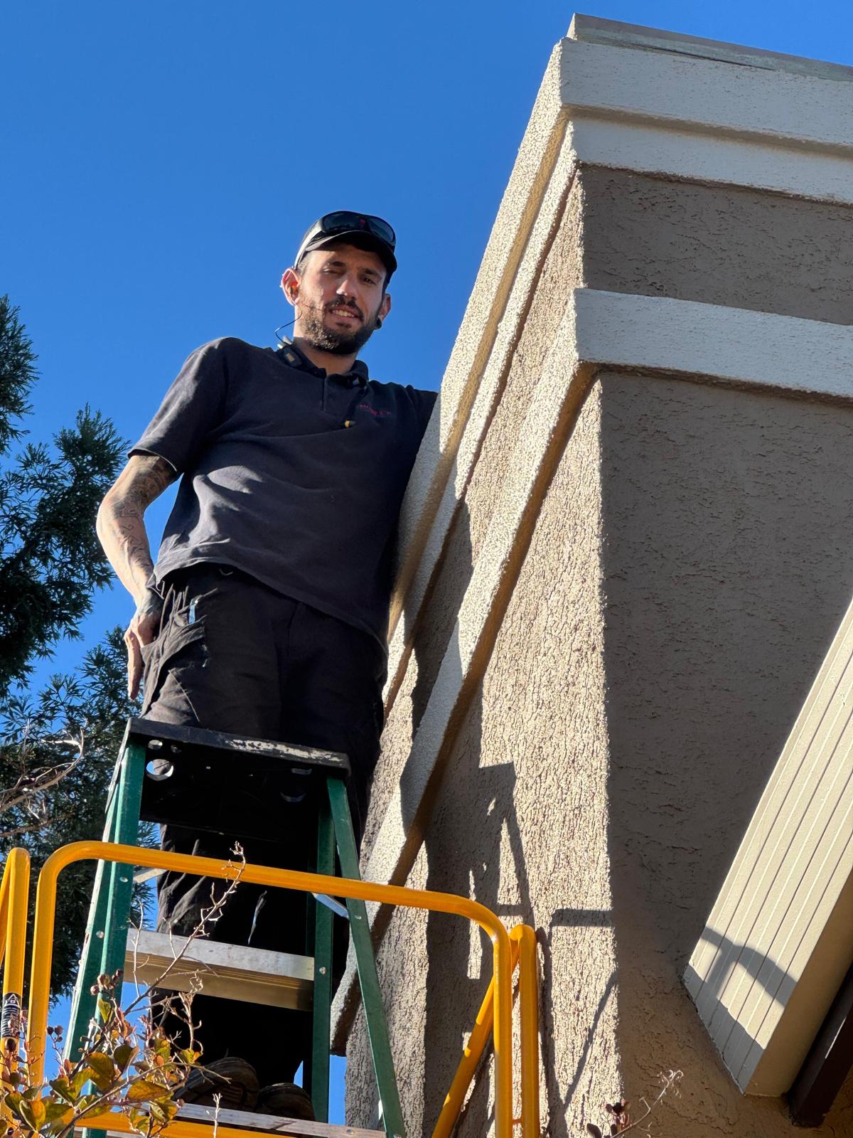 Michael near the top of a green step ladder beside a tall stucco chimney
