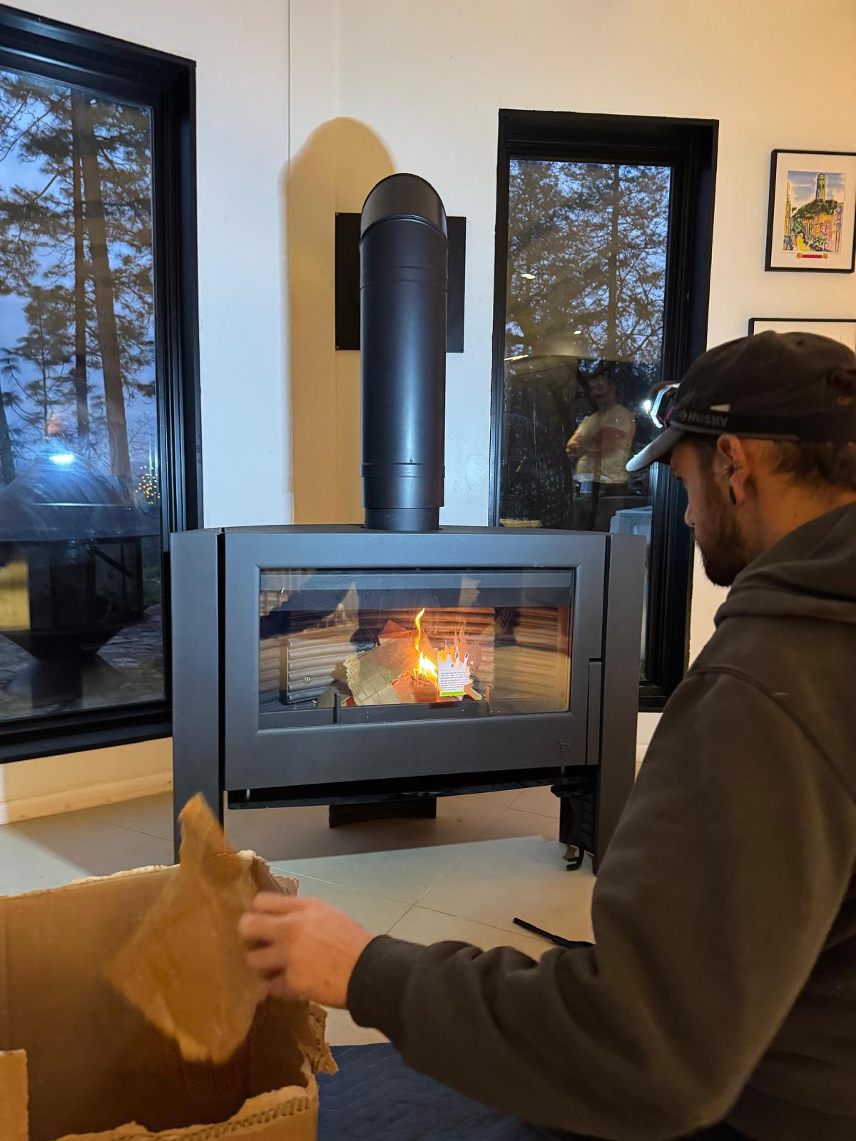 A fire burning behind the glass of a black stove as Michael reaches into a cardboard box